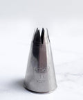 A stainless steel 1M piping tip with an open star design, standing upright on a light gray marble surface against a white background.