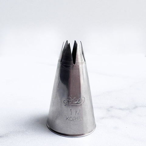 A stainless steel 1M piping tip with an open star design, standing upright on a light gray marble surface against a white background.