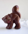 A 3d cat made from milk chocolate with green eyes and a red mouth, with it's tail up in the air. Standing along side the chocolate mold used to make it.