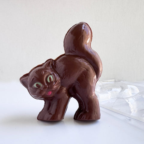 A 3d cat made from milk chocolate with green eyes and a red mouth, with it's tail up in the air. Standing along side the chocolate mold used to make it.