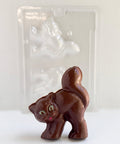 A 3d chocolate cat made from milk chocolate with green eyes and a red mouth with it's tail up in the air. Placed in front of a plastic chocolate mold that was used to make it.