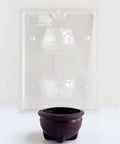 A small dark brown cauldron-shaped chocolate shown in front of a clear plastic candy mold tray on a plain white background.