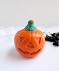 An orange jack-o-lantern-shaped 3d chocolate with a green stem, photographed on a white surface with a clear plastic chocolate mold tray behind it and black binder clips to the right.