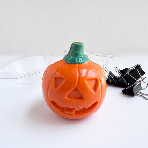 An orange jack-o-lantern-shaped 3d chocolate with a green stem, photographed on a white surface with a clear plastic chocolate mold tray behind it and black binder clips to the right.