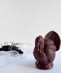 Dark chocolate 3d molded turkey with fine detail along side the chocolate mold that is cut for molding and the binder clips sitting next to it that are used to secure the mold while filled with chocolate.