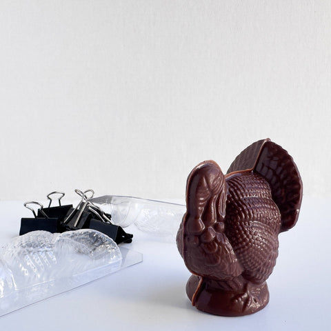 Dark chocolate 3d molded turkey with fine detail along side the chocolate mold that is cut for molding and the binder clips sitting next to it that are used to secure the mold while filled with chocolate.