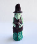 A 3d chocolate witch with pale green skin, wearing a dark brown hat and dress, and a green textured base, photographed against a plain white background.