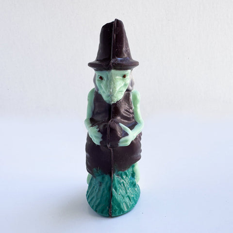 A 3d chocolate witch with pale green skin, wearing a dark brown hat and dress, and a green textured base, photographed against a plain white background.
