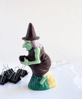 A small 3d chocolate witch with a green face and hands, wearing a dark chocolate hat and dress and riding a broom. The chocolate witch is photographed on a white surface with black binder clips on the left and a clear plastic chocolate mold on the right against a white background.