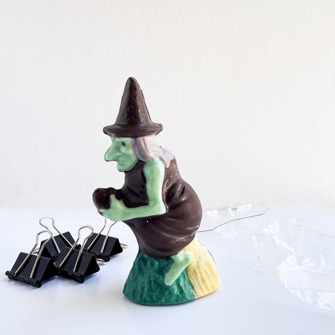 A small 3d chocolate witch with a green face and hands, wearing a dark chocolate hat and dress and riding a broom. The chocolate witch is photographed on a white surface with black binder clips on the left and a clear plastic chocolate mold on the right against a white background.