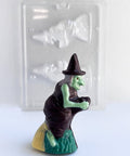 A small witch 3d chocolate with a green face and legs, wearing a dark chocolate hat and dress, riding a broomstick with a yellow and green base, shown in front of a clear plastic candy mold.