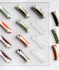 A clear plastic bone-shaped candy mold or tray with multiple cavities, shown on a white background. The cavities contain small bone candies in white, dark brown, green, and orange chocolate, while other cavities are empty and clear.