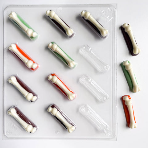 A clear plastic bone-shaped candy mold or tray with multiple cavities, shown on a white background. The cavities contain small bone candies in white, dark brown, green, and orange chocolate, while other cavities are empty and clear.