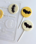 A clear plastic round lollipop mold with a single bat molded in the center on a white surface beside two decorated lollipops