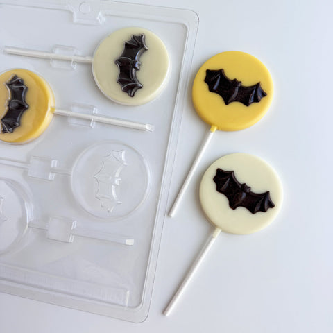 A clear plastic round lollipop mold with a single bat molded in the center on a white surface beside two decorated lollipops