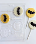 A clear plastic Halloween-themed bat lollipop candy mold shown next to two finished lollipops decorated with black bat shapes on yellow and white backgrounds.