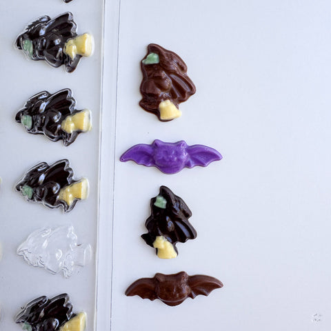 A Halloween chocolate mold and finished chocolate pieces shaped like small witches and bats, shown on a white background.