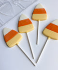 Four candy corn shaped lollipops with white, orange, and yellow stripes on white sticks, arranged on a white surface next to a clear plastic mold.
