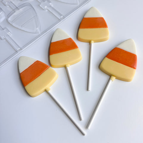 Four candy corn shaped lollipops with white, orange, and yellow stripes on white sticks, arranged on a white surface next to a clear plastic mold.