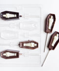 A Halloween-themed lollipop mold featuring coffin-shaped molds with mummy figures inside, shown alongside two finished dark chocolate and white chocolate lollipops on a clean white background.