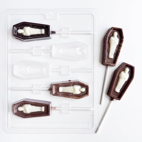 A Halloween-themed lollipop mold featuring coffin-shaped molds with mummy figures inside, shown alongside two finished dark chocolate and white chocolate lollipops on a clean white background.