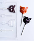 A clear plastic lollipop mold tray on a white background, shown next to two finished cat-face chocolate lollipops in orange and dark chocolate with white lollipop sticks.