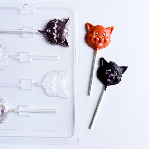 A clear plastic lollipop mold tray on a white background, shown next to two finished cat-face chocolate lollipops in orange and dark chocolate with white lollipop sticks.