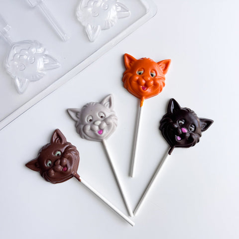 Four cat-face lollipops in dark chocolate, gray, orange, and milk chocolate, arranged on a white surface with a clear plastic candy mold partially visible in the upper left corner.