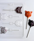 A clear plastic candy mold with four cat-face cavities shown next to three finished cat-shaped lollipops in dark chocolate, orange chocolate, and gray chocolate, all on a white background.