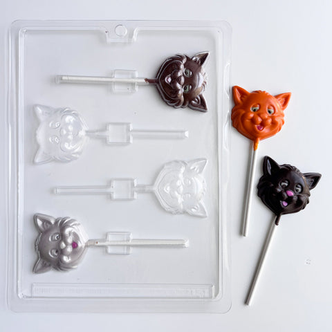 A clear plastic candy mold with four cat-face cavities shown next to three finished cat-shaped lollipops in dark chocolate, orange chocolate, and gray chocolate, all on a white background.