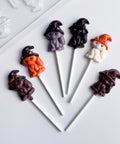 6 chocolate lollipops shaped like cats with witches hats on their heads. Each lollipops is made with different colored chocolate, and positioned next to the clear plastic candy mold