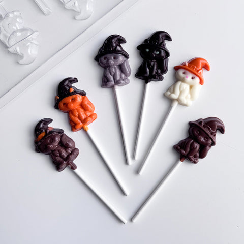 6 chocolate lollipops shaped like cats with witches hats on their heads. Each lollipops is made with different colored chocolate, and positioned next to the clear plastic candy mold