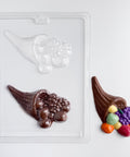 A clear plastic candy mold tray showing two cornucopia-shaped cavities, one empty and one filled with dark brown chocolate. A finished chocolate cornucopia with colorful  fruits spilling out sits to the right on a clean white background.