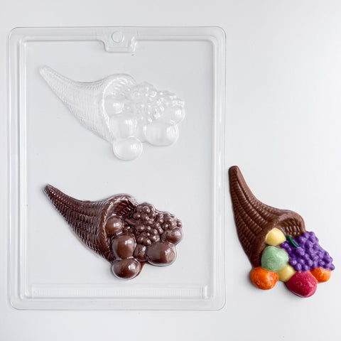 A clear plastic candy mold tray showing two cornucopia-shaped cavities, one empty and one filled with dark brown chocolate. A finished chocolate cornucopia with colorful  fruits spilling out sits to the right on a clean white background.