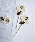 Two decorated white chocolate lollipops shaped like smiling ghosts with small dark brown bat details, shown on a white background next to a clear plastic mold.