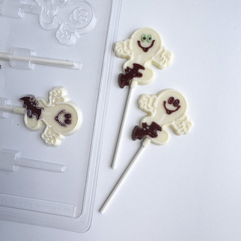 Two decorated white chocolate lollipops shaped like smiling ghosts with small dark brown bat details, shown on a white background next to a clear plastic mold.