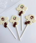 Four white chocolate ghost lollipops with dark brown chocolate details and white sticks, arranged on a white background.