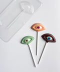 Three novelty eye-shaped lollipops with white lollipop sticks are arranged on a white surface next to a clear plastic chocolate mold. The eyes are colored green, peach, and dark brown, each with a small realistic pupil detail.