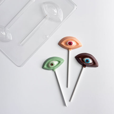 Three novelty eye-shaped lollipops with white lollipop sticks are arranged on a white surface next to a clear plastic chocolate mold. The eyes are colored green, peach, and dark brown, each with a small realistic pupil detail.
