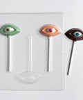 human eye shaped chocolate lollipops with upper and lower eye lid and eyeball along side the plastic candy mold to make them sitting on a white background. The chocolate eyes are made in a variety of colors like green colored chocolate, flesh color, and dark chocolate.