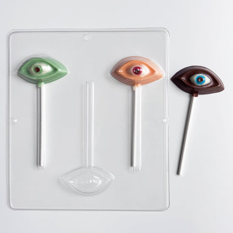 human eye shaped chocolate lollipops with upper and lower eye lid and eyeball along side the plastic candy mold to make them sitting on a white background. The chocolate eyes are made in a variety of colors like green colored chocolate, flesh color, and dark chocolate.