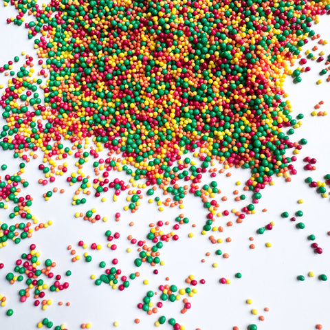 Multicolor round ball shaped nonpareil sprinkles in red, green, yellow, and orange scattered on a white background, with a larger pile concentrated toward the top right.