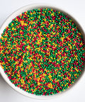 A top-down view of a white bowl filled with small, round, fall colored nonpareil sprinkles in green, red, orange, and yellow on a white background.