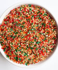 A top-down view of a white bowl filled with a fall colored mix of coarse sugar crystal sprinkles in orange, green, red, yellow, and metallic gold, sitting on a white back drop.