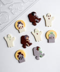 A set of Halloween-themed chocolate decorations and a clear plastic candy mold  on a white background. The decorations include ghost shapes, zombie figures in brown and gray, tombstones with 'RIP' text, and a demon on a yellow circle.