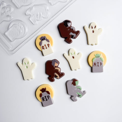 A set of Halloween-themed chocolate decorations and a clear plastic candy mold  on a white background. The decorations include ghost shapes, zombie figures in brown and gray, tombstones with 'RIP' text, and a demon on a yellow circle.