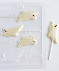 White chocolate ghost-shaped lollipops shown next to a clear plastic ghost chocolate mold and one finished ghost lollipop on a white background.