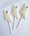Three ghost-shaped lollipops made with white chocolate with small dark brown eyes, displayed on a white surface.