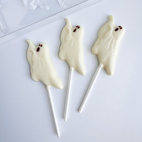 Three ghost-shaped lollipops made with white chocolate with small dark brown eyes, displayed on a white surface.