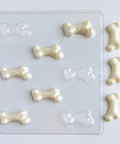 A clear plastic bone-shaped candy mold with several white chocolate bone pieces arranged beside it on a plain white background.